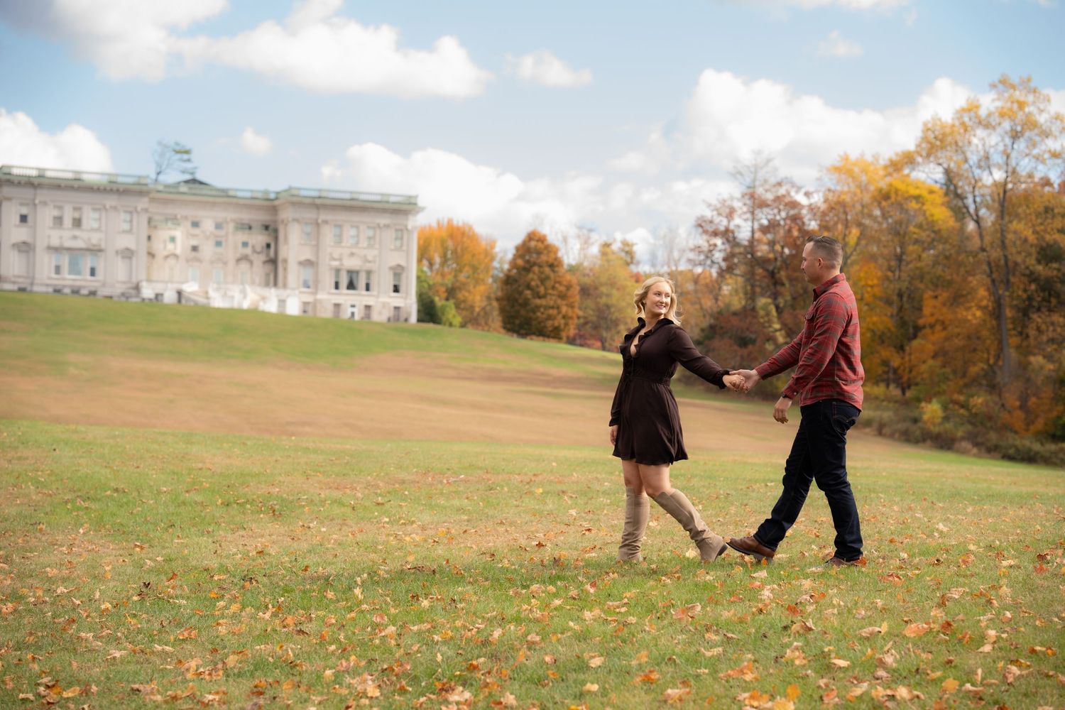 Couple holding hands walking on green grass field with colorful autumn trees and grand historic building in background.