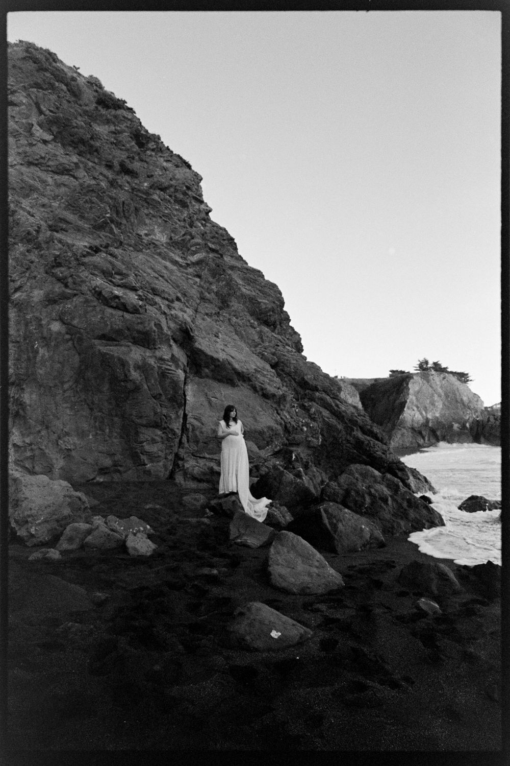 Pregnant woman in flowing white gown standing on rocky coastal cliffs by the ocean in black and white photo.