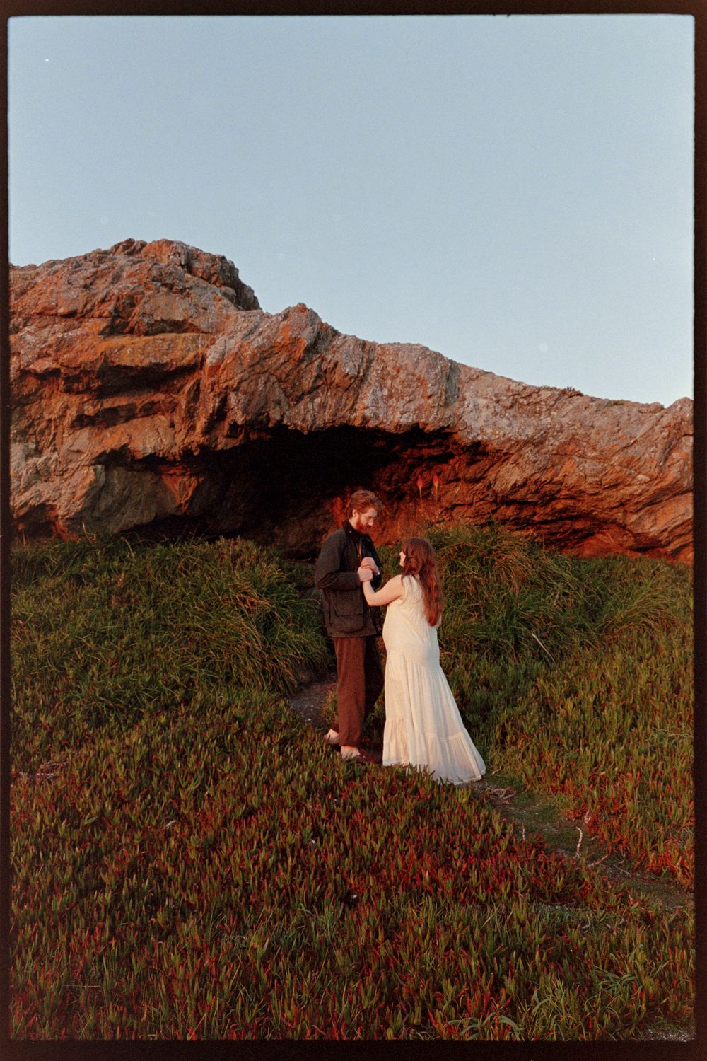 A couple in wedding attire share a romantic moment in front of dramatic coastal rocks at golden hour.