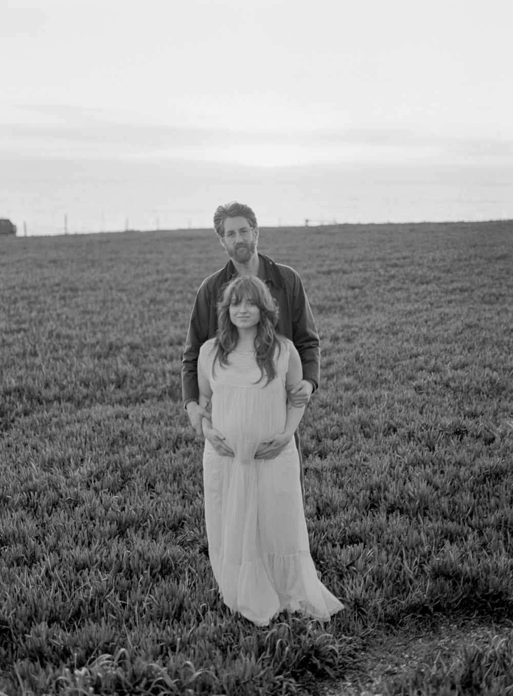 Couple poses in open field at dusk, man embracing pregnant woman in white dress.