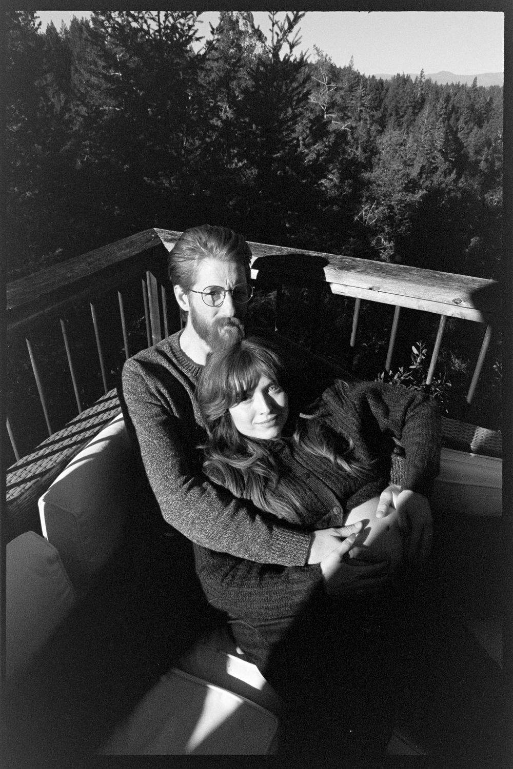 A couple embraces tenderly on a wooden deck surrounded by forest trees in a black and white portrait photo.