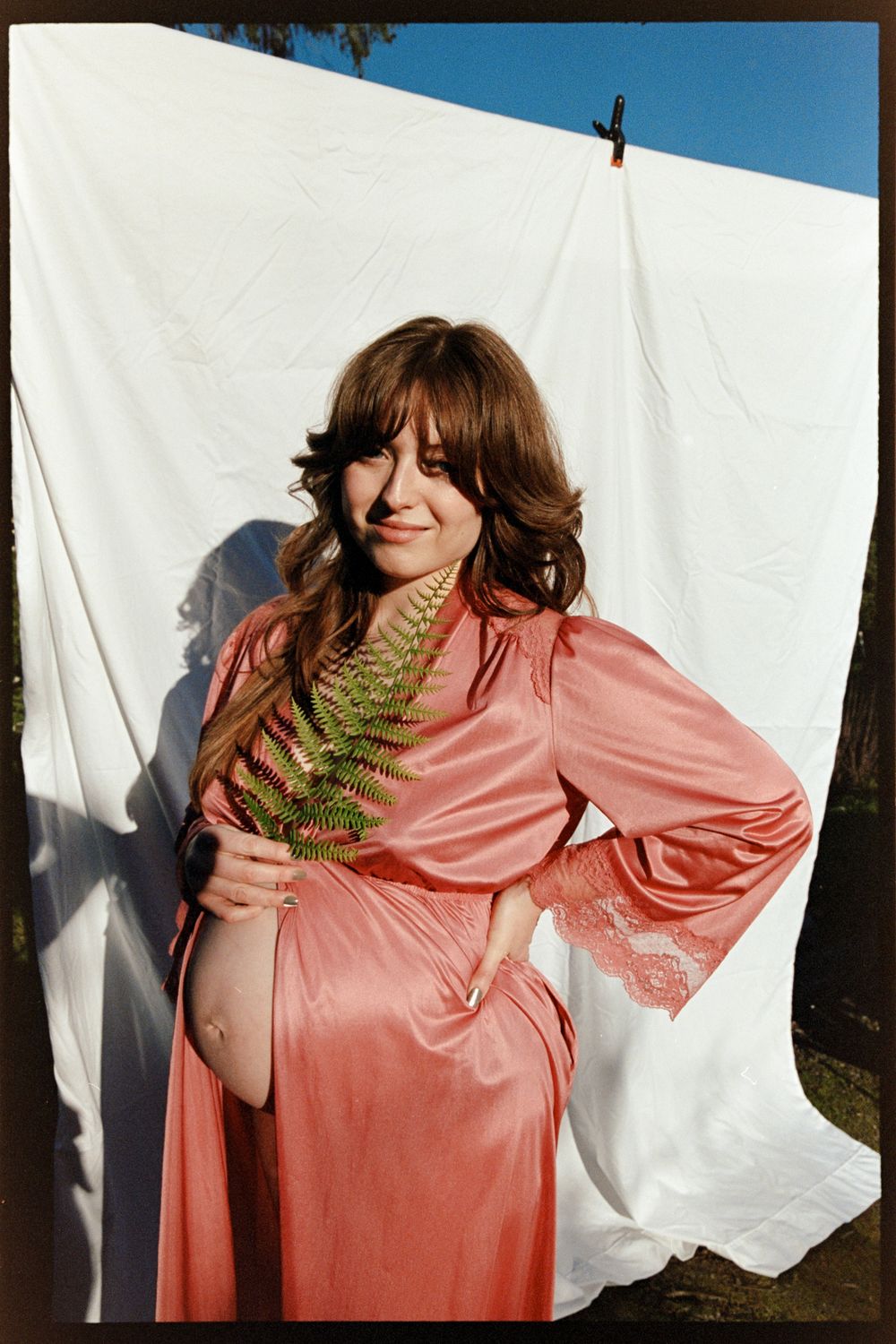 Pregnant woman in pink satin robe holds fern outdoors against white sheet backdrop.