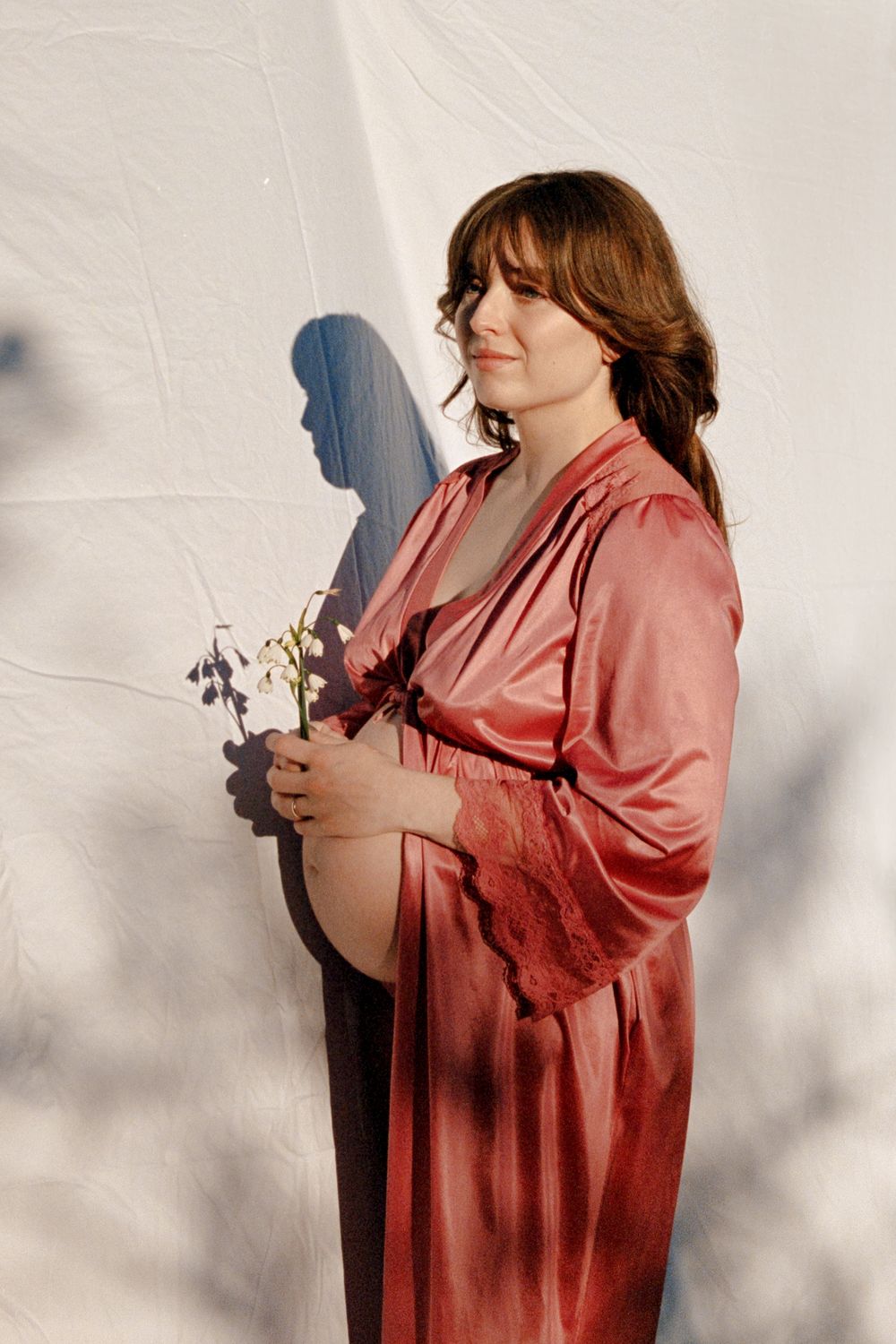 Pregnant woman in pink satin robe holding white flowers against white backdrop.