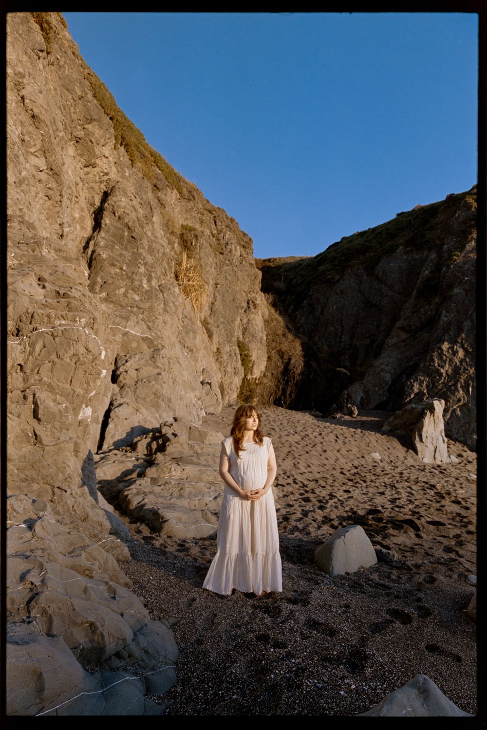 A woman in a white dress stands on a rocky beach surrounded by towering cliffs under a clear blue sky.
