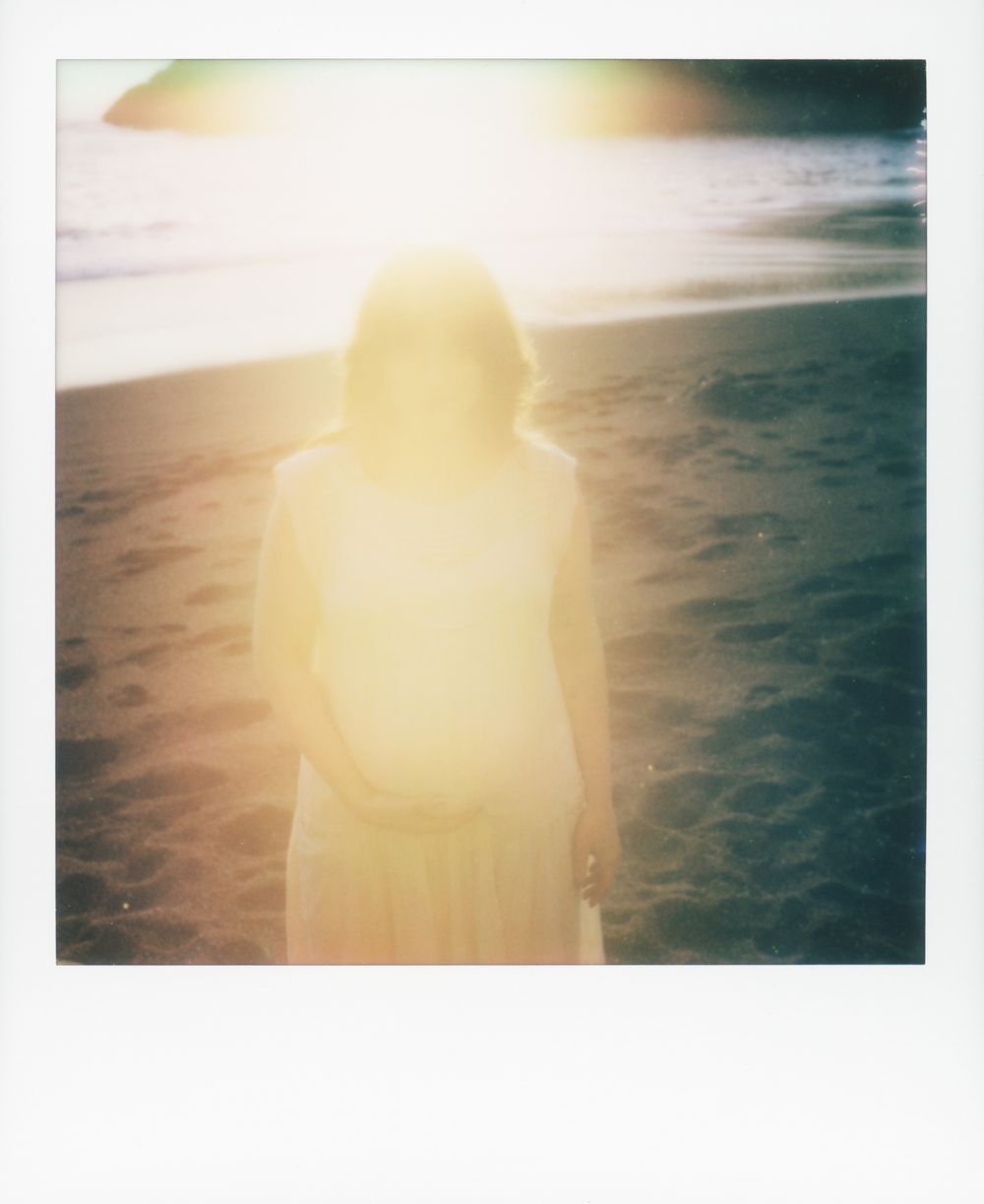 A woman in a white dress stands on a beach, backlit by bright sunlight creating a dreamy overexposed glow.