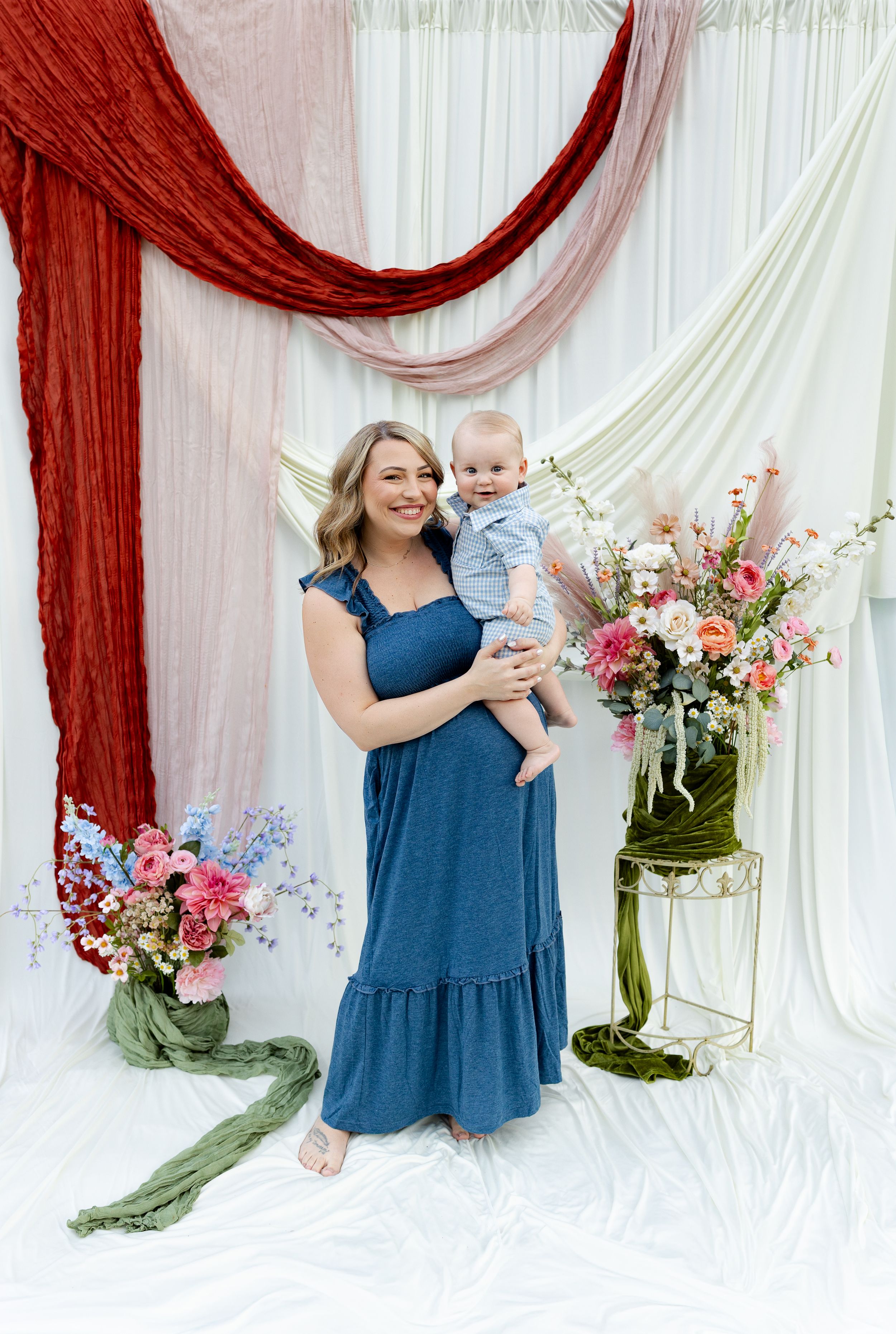 A smiling woman in a blue maxi dress holds a baby boy, posed before draped fabric and floral arrangements.