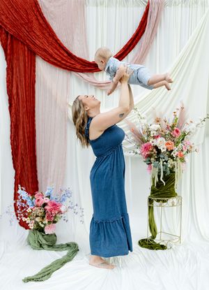 A woman in a blue maxi dress lifts her baby overhead, surrounded by colorful floral arrangements and draped fabric backdrops.