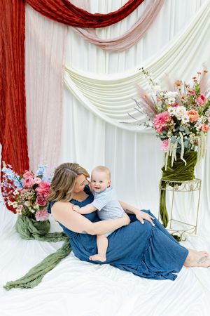 A mother in a blue dress sits on white fabric holding her baby boy, surrounded by colorful floral arrangements and draped fabric.