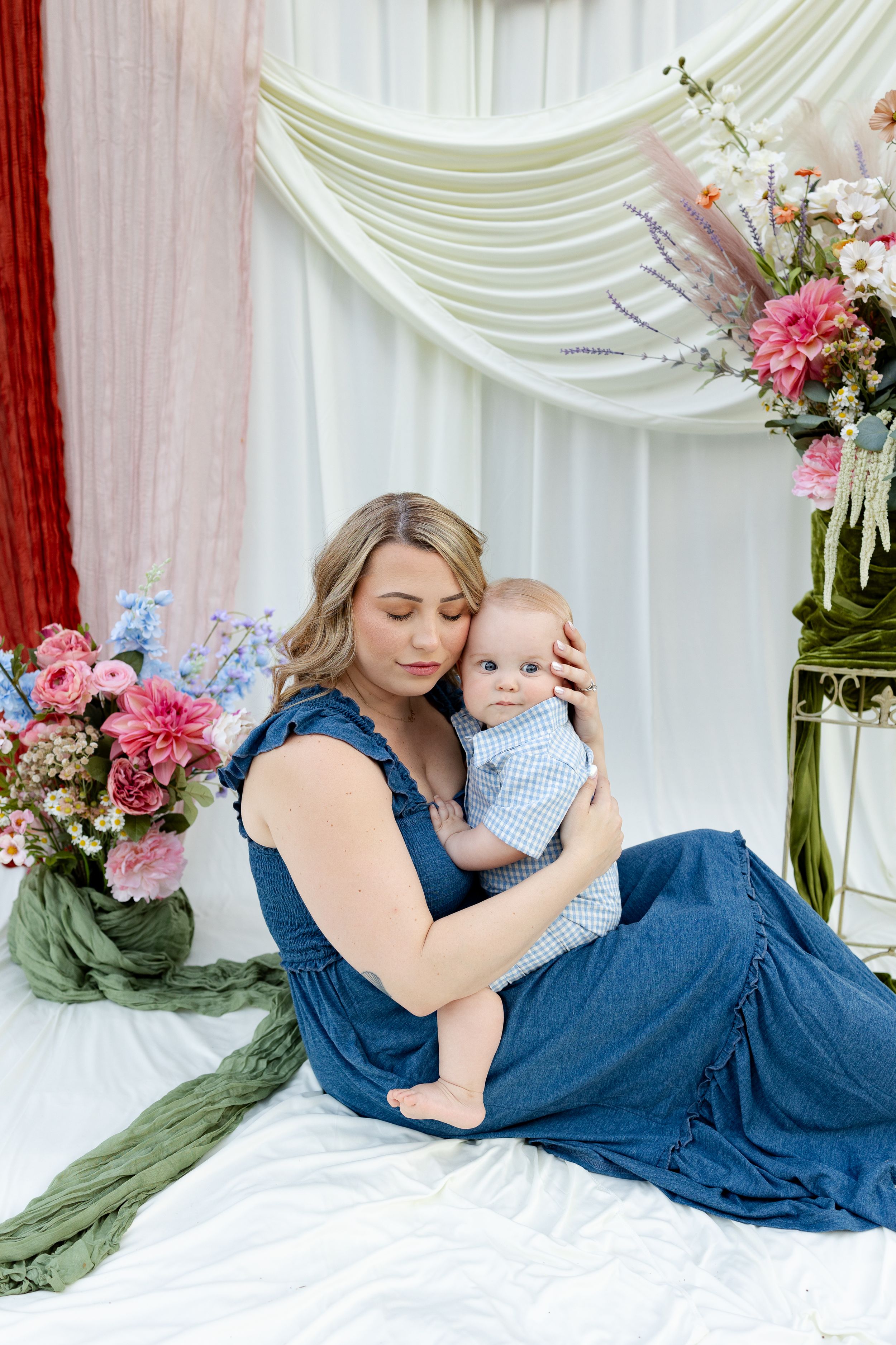 A mother in a blue dress tenderly holds her baby boy amid colorful floral arrangements and draped fabric backdrop.