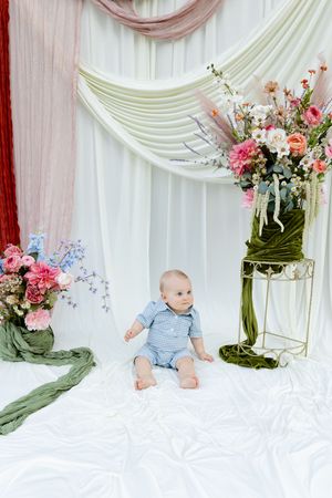 Adorable baby boy in blue romper sits alone surrounded by lush floral arrangements and elegant draped fabric backdrop.