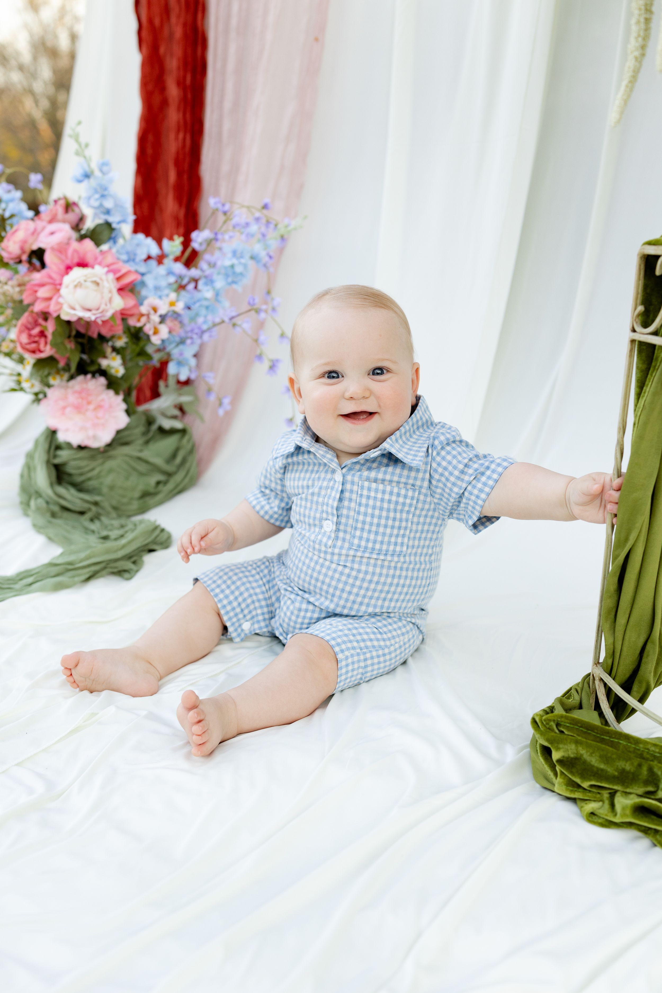 Smiling baby boy in blue gingham romper sitting on white fabric surrounded by colorful spring flowers.