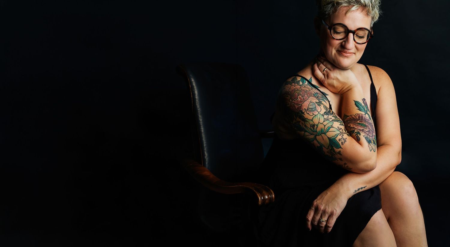 Smiling woman with colorful tattoos on her arm sitting on a leather chair against a dark background.
