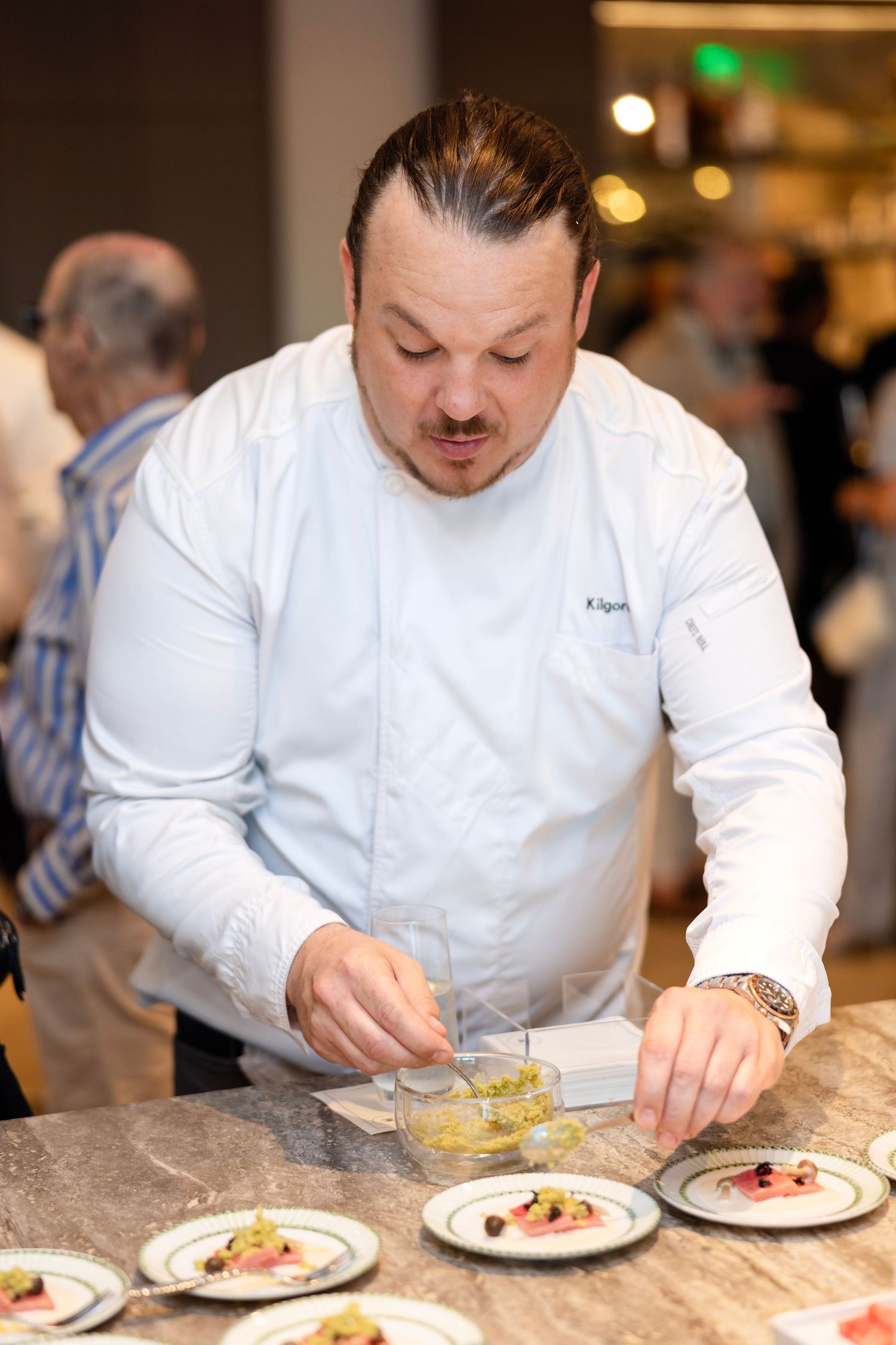 Chef in white uniform carefully plating elegant dishes at a marble counter during a culinary event.