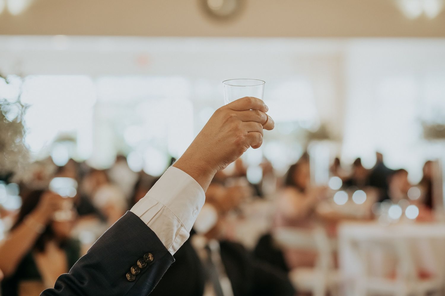A man in a suit raises a glass toast at an elegant wedding reception venue filled with guests.
