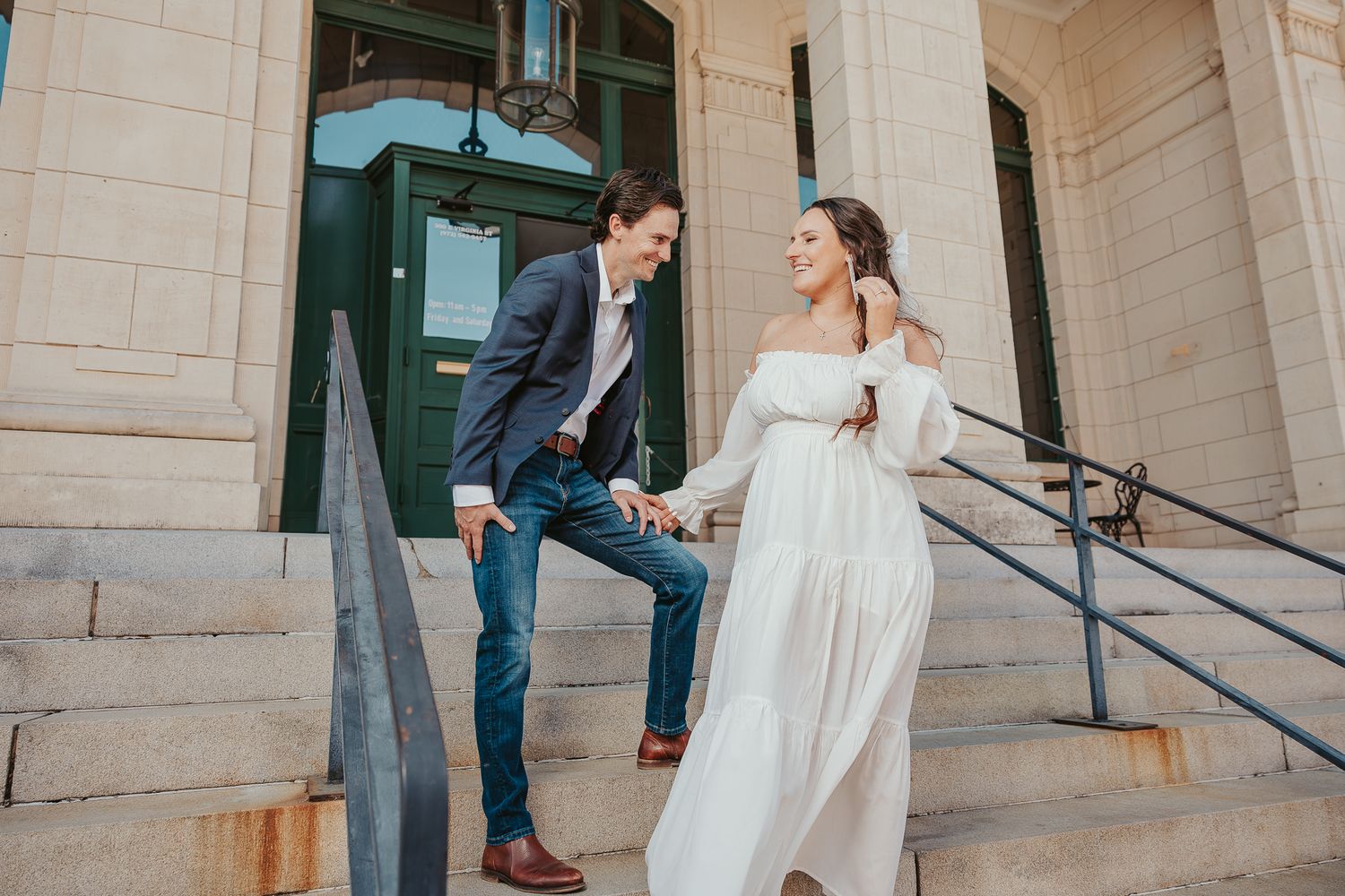 Couple holding hands walking down stone steps, woman in white off-shoulder gown, man in blue blazer and jeans.