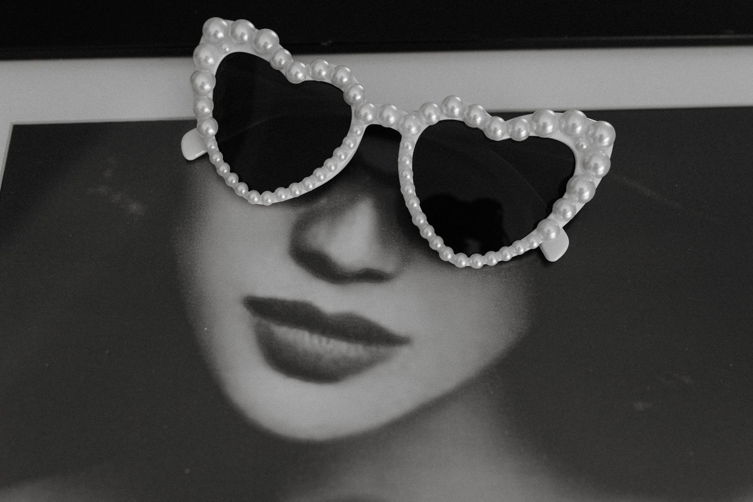 Heart-shaped pearl sunglasses resting on a black and white portrait photo of a glamorous woman.