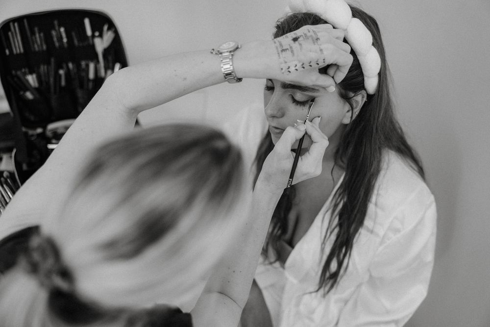 Bride getting makeup applied by makeup artist while wearing white robe and headband on wedding day.
