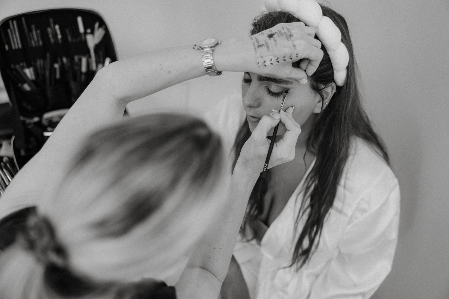 Bride getting makeup applied by makeup artist while wearing white robe and headband on wedding day.