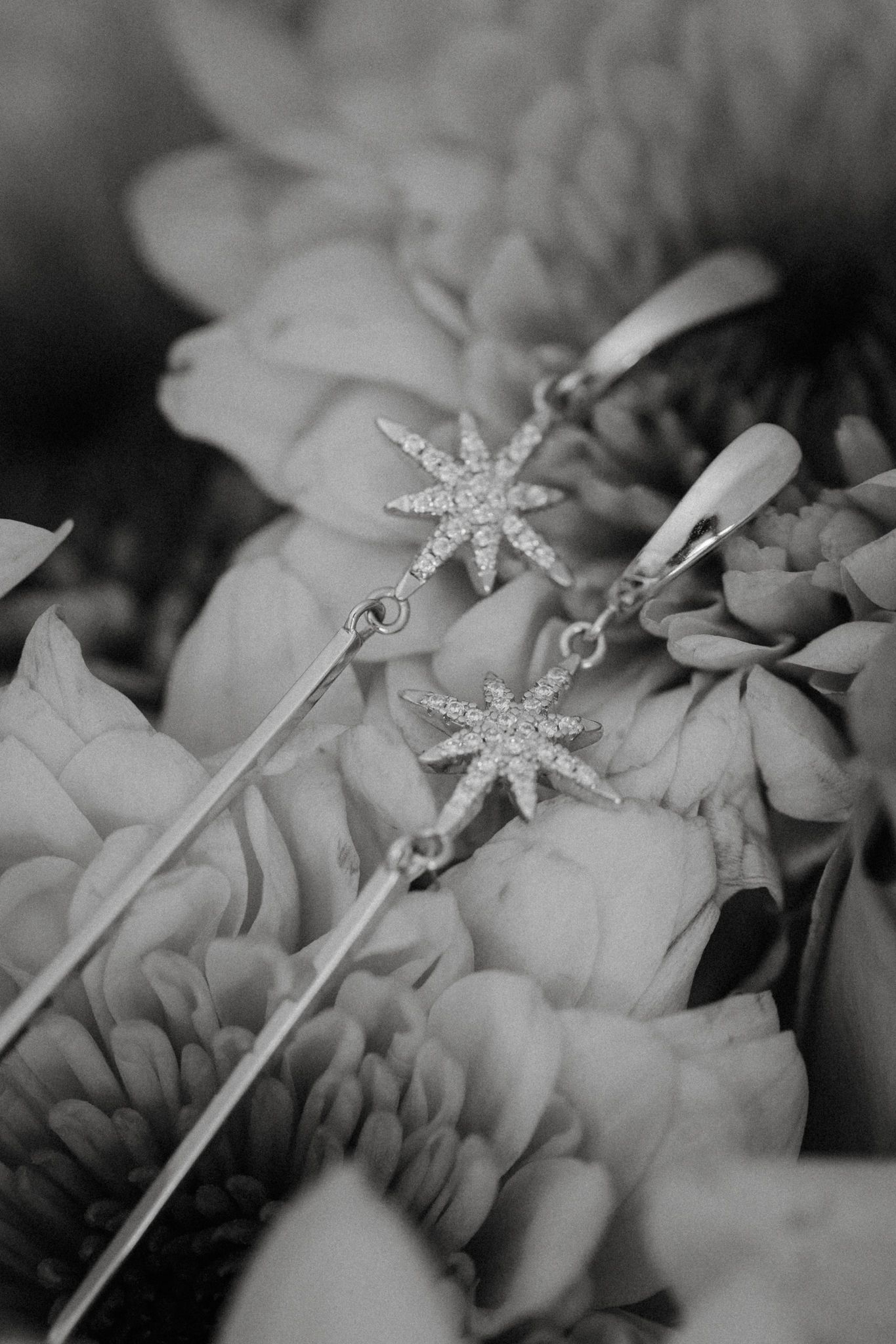 Black and white photo of elegant star-shaped diamond drop earrings resting on a bed of fresh flowers.