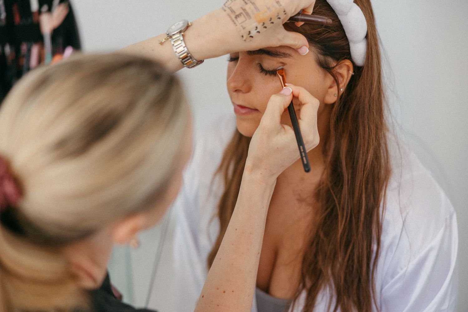 A makeup artist applying eye makeup to a woman with long brown hair using a brush in a bright studio.