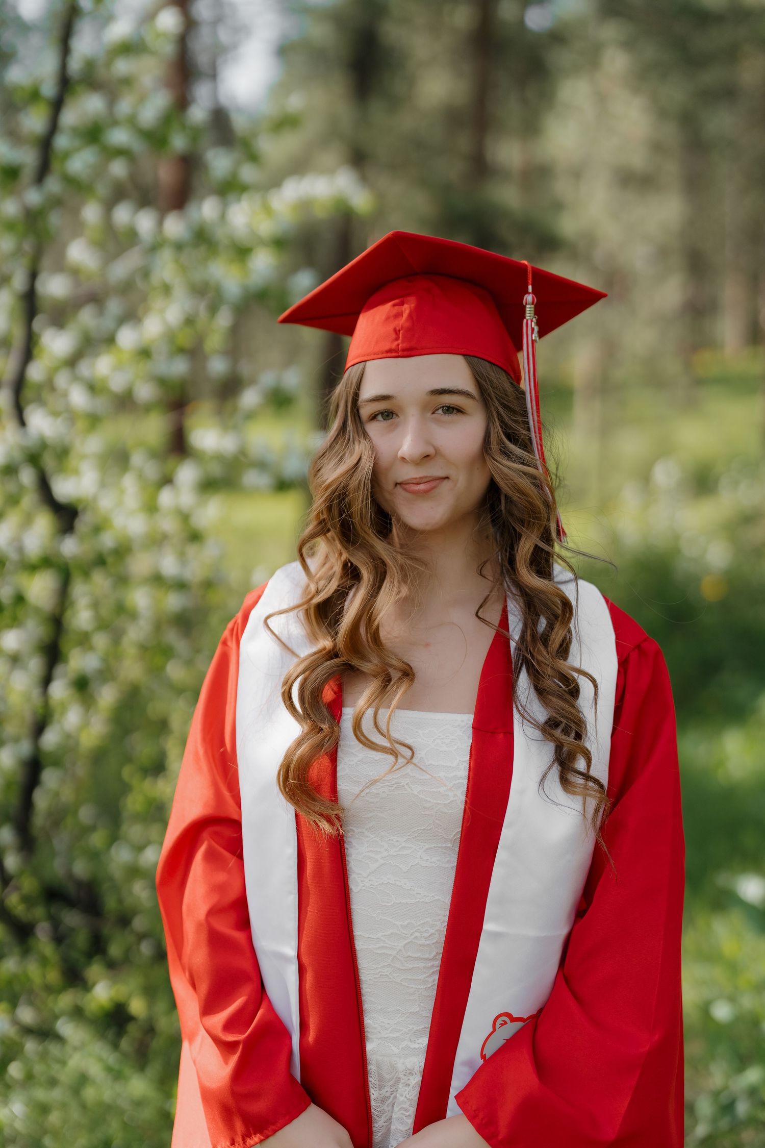 Young woman in red graduation cap and gown smiling outdoors among blooming trees.