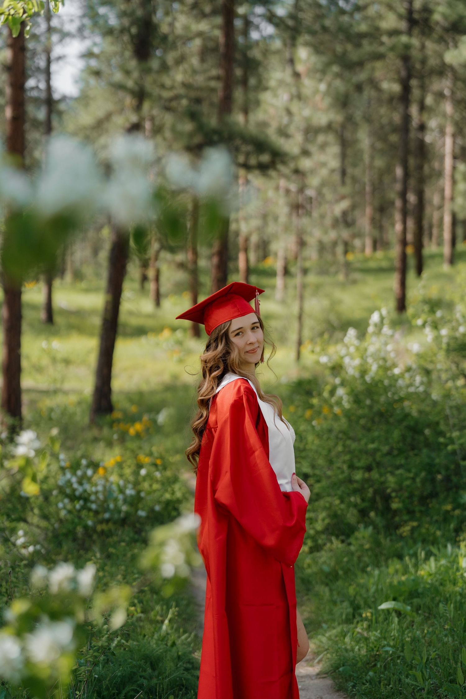Young woman in red graduation cap and gown poses outdoors in a lush green forest setting.