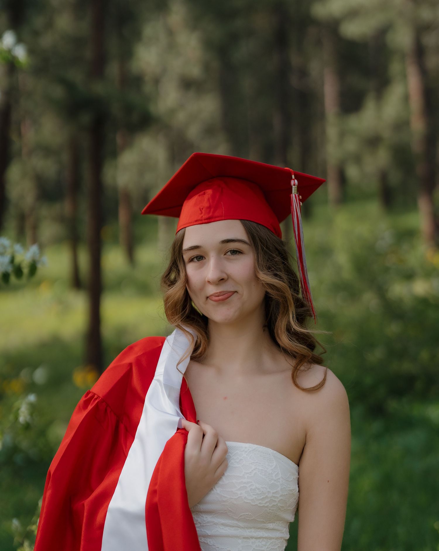 Young woman in red graduation cap and gown smiling outdoors in a lush green forest setting.