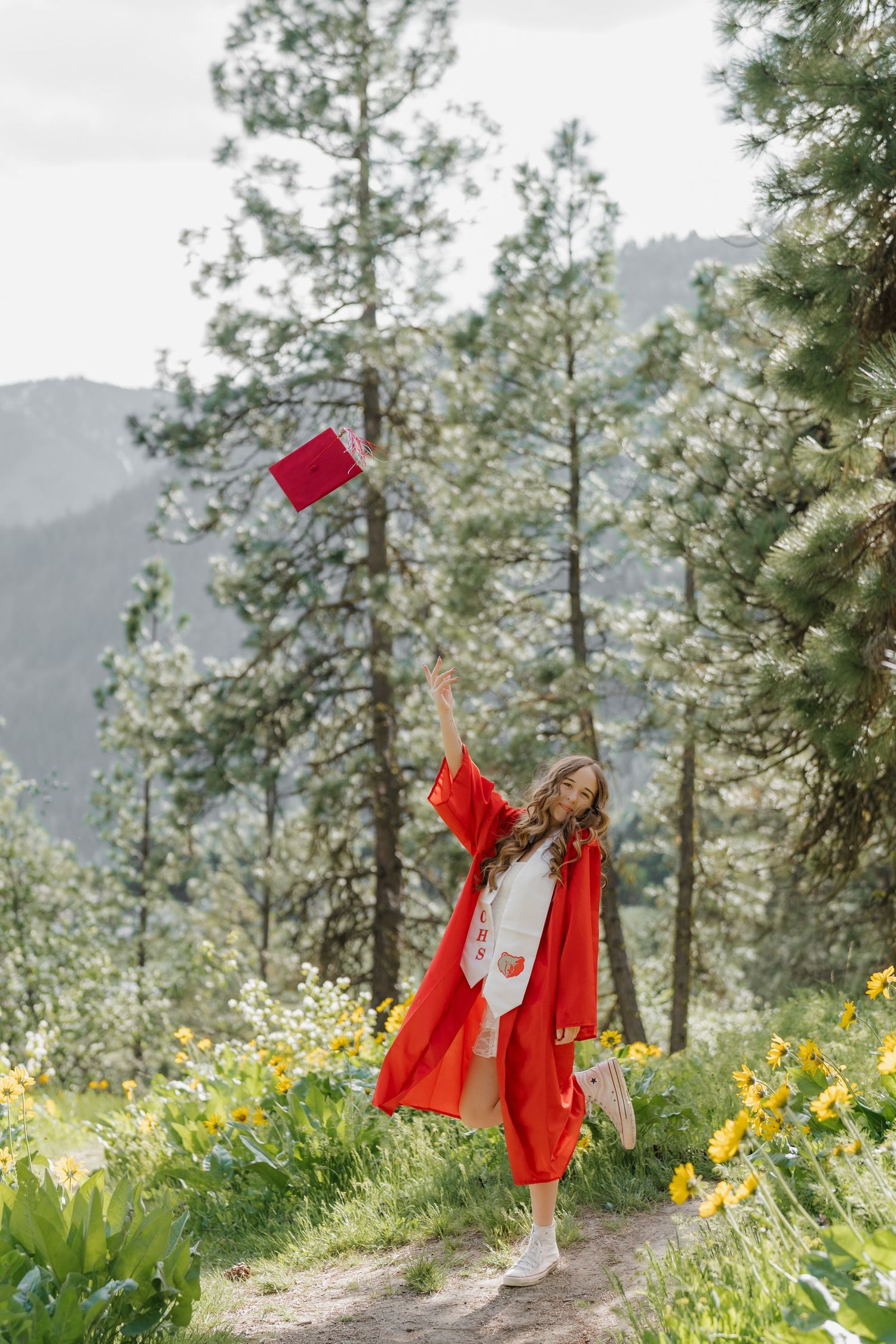 Graduate in red gown tosses cap in mountain meadow with wildflowers and pine trees.