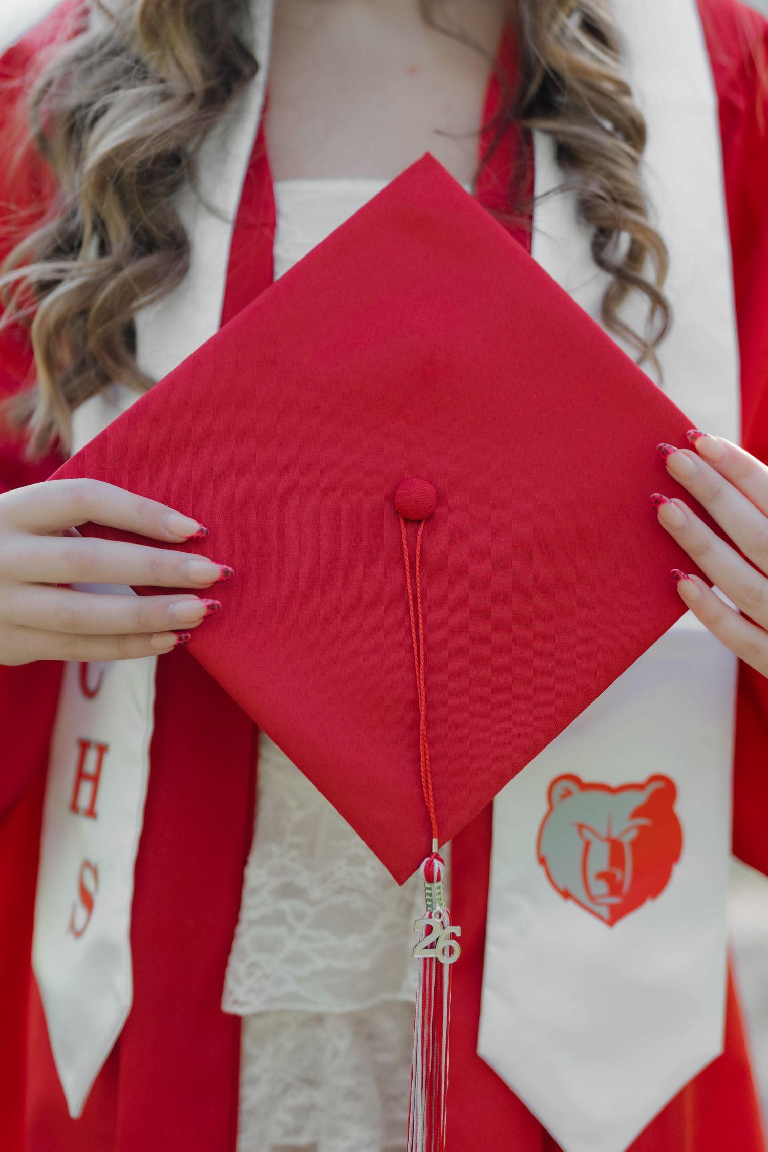 Student in red graduation gown holding red mortarboard cap with bear mascot tassel charm.
