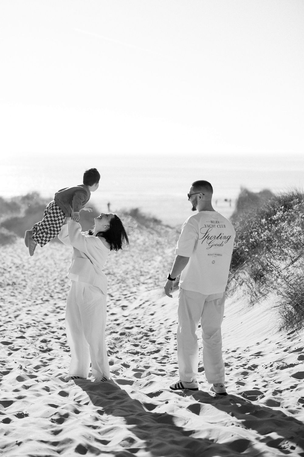 Black and white photo of a family on sandy beach dunes, mother lifting baby while father watches, ocean in background.