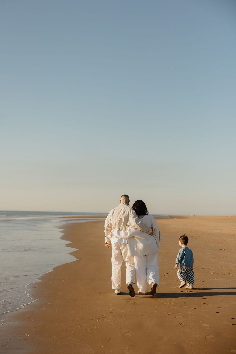 Family of three walking along a sandy beach at sunset, parents in white clothing with young child in blue outfit.
