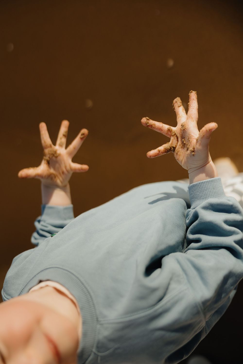 Child in blue sweatshirt holding up two muddy hands with fingers spread wide against a dark brown background.