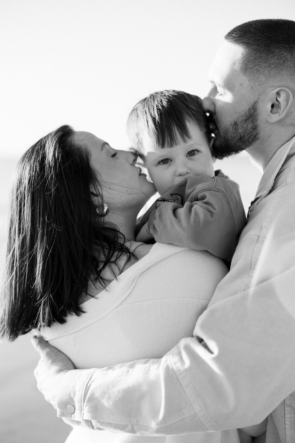 Black and white photo of parents kissing their baby on the cheeks, sharing a tender family moment.