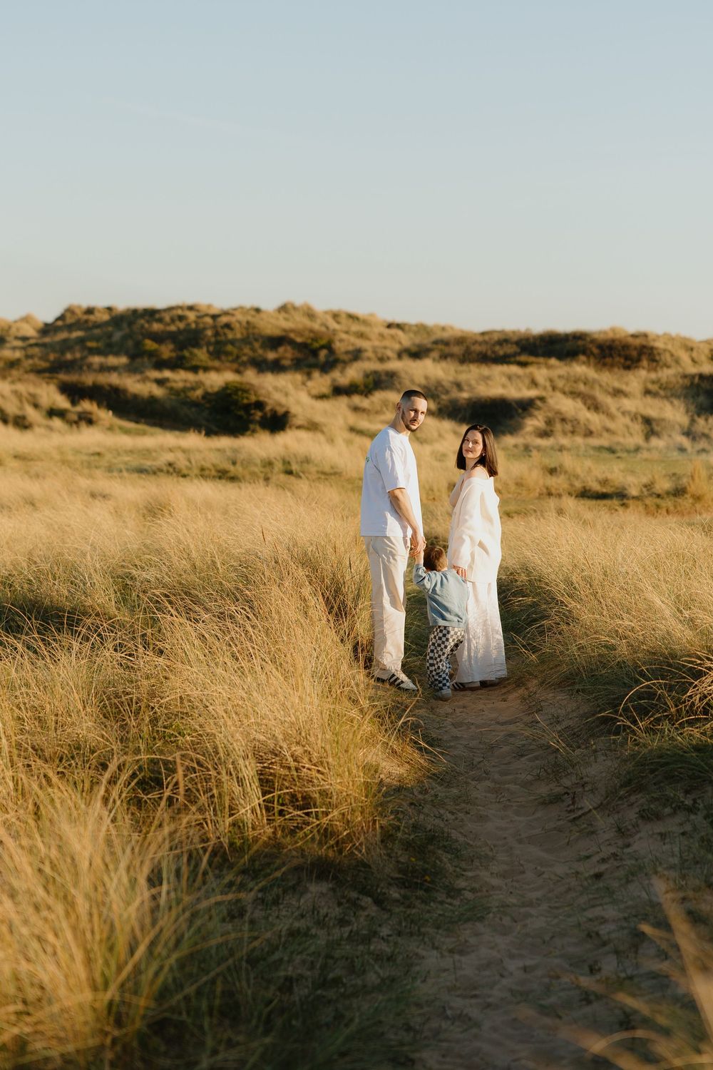 Family in white outfits walking on a sandy path through golden grass dunes at sunset.