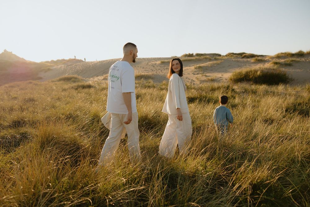 Family of three walking through golden grass dunes at sunset, wearing white outfits in a serene coastal landscape.