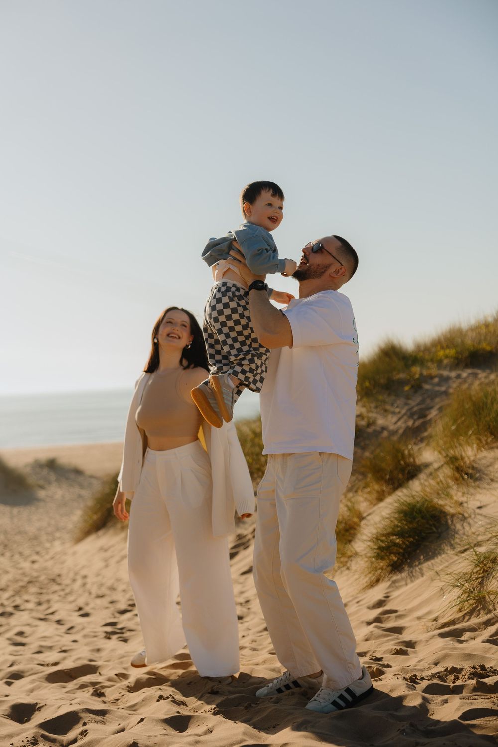 Happy family on sandy beach dunes, parents lifting smiling baby in the air during golden hour sunset.
