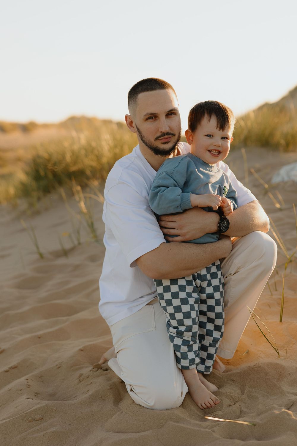 Father kneeling on sandy dunes holding smiling toddler, both wearing casual clothes in warm golden sunset light.