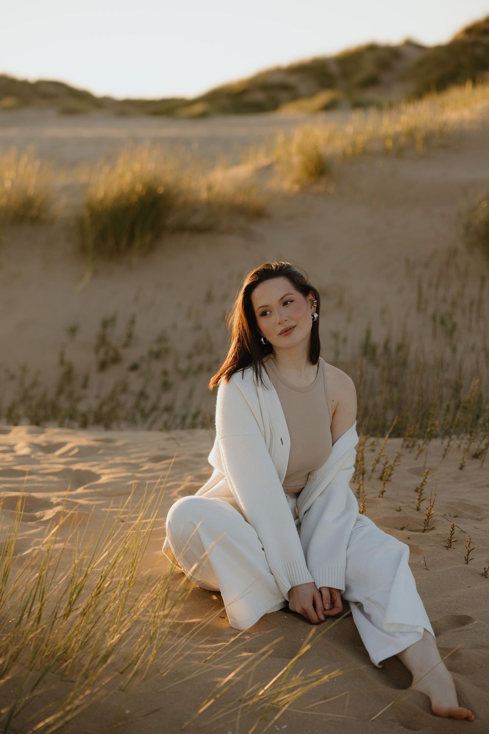 A young woman in white linen outfit sits barefoot on golden sand dunes at sunset.