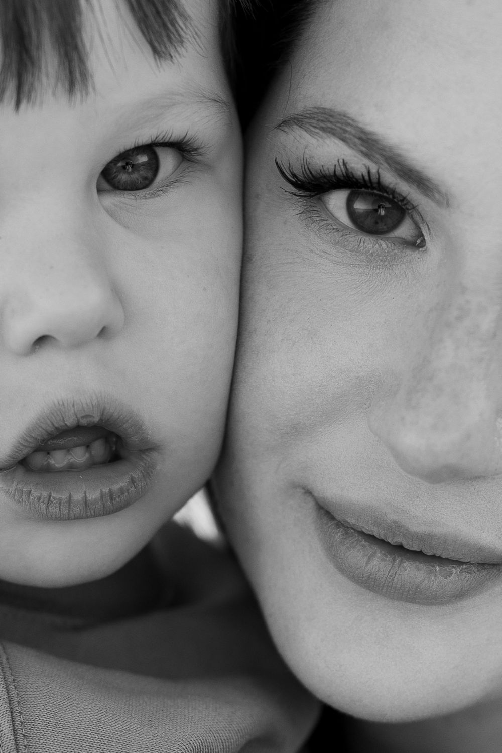 Black and white close-up portrait of a young child and smiling woman cheek to cheek, showing expressive eyes.