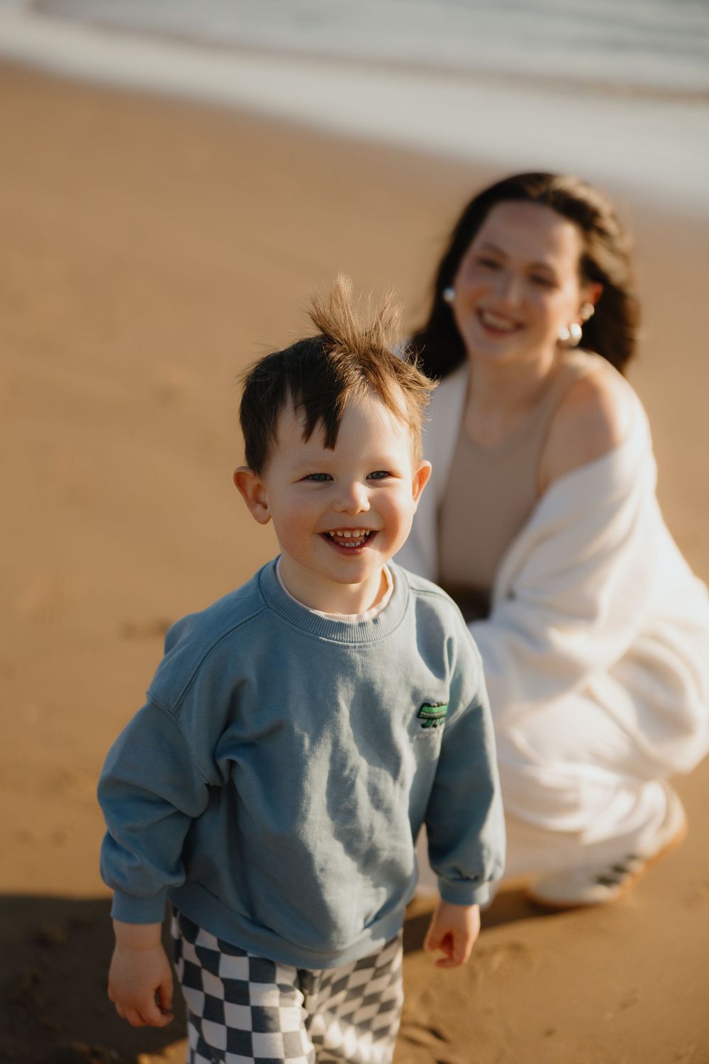 Smiling toddler boy in blue sweatshirt and checkered shorts plays on sandy beach with mother in background.