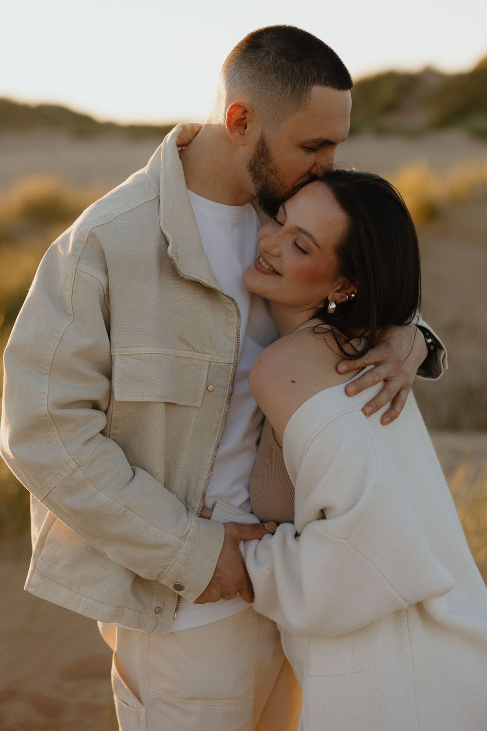 Couple embracing at golden hour on sandy dunes, woman smiling as man kisses her head, both wearing neutral tones.