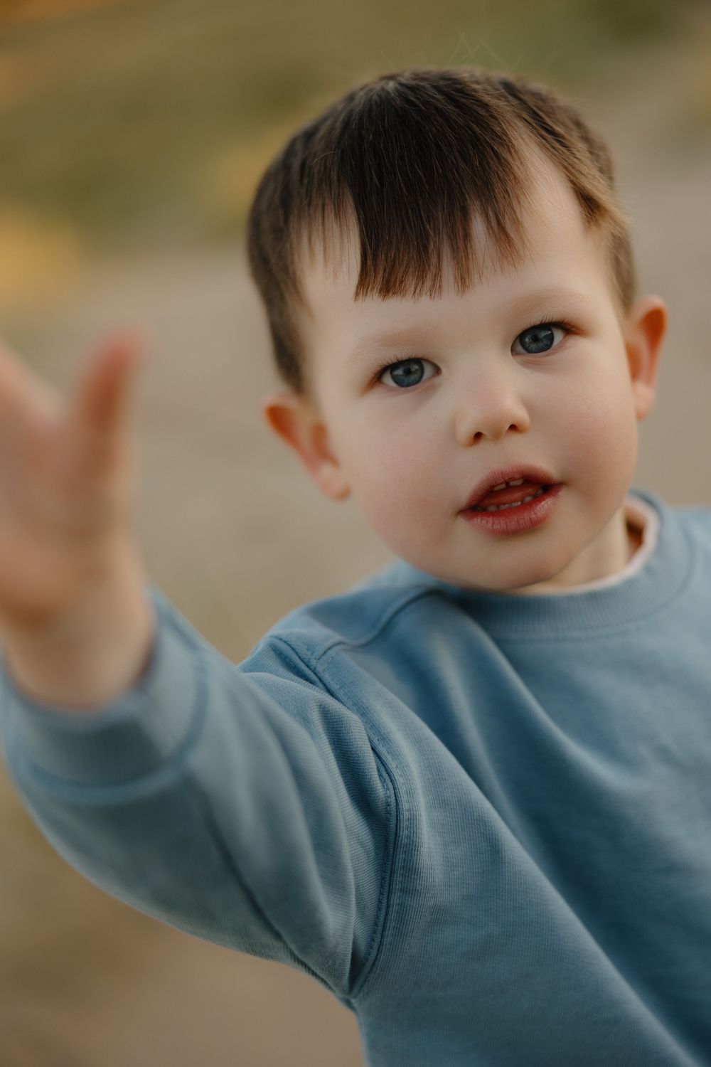 Cute toddler boy in a blue sweatshirt reaching his hand toward the camera outdoors.