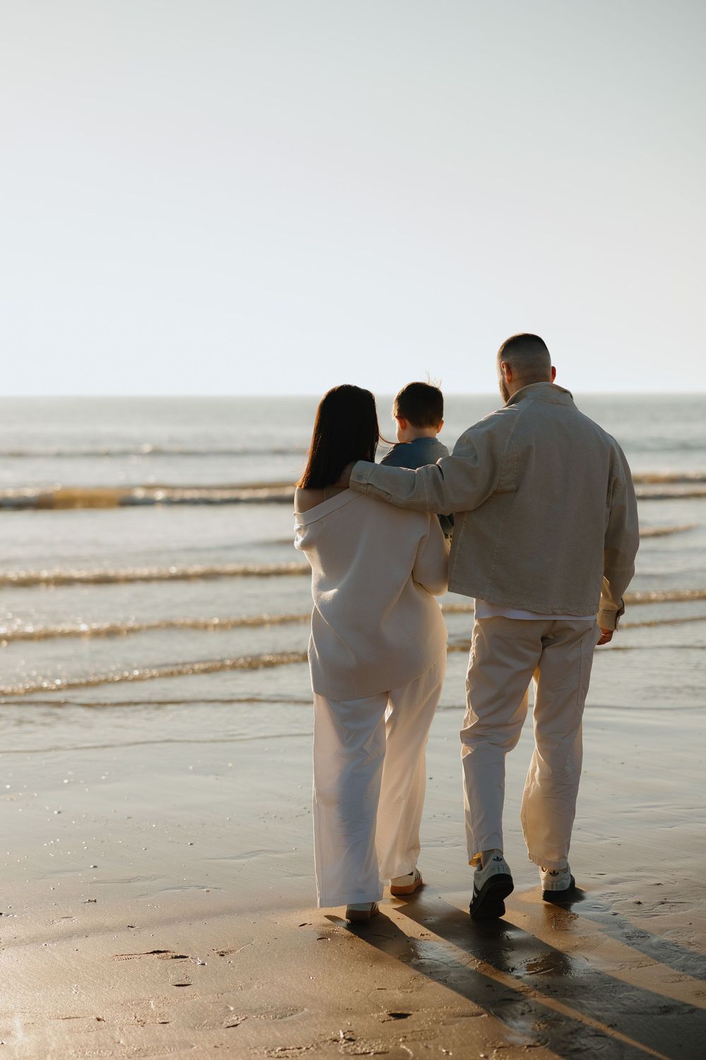 Family of three walking on a sandy beach at sunset, viewed from behind, wearing light casual clothing.