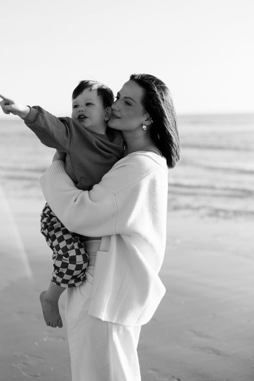 Black and white photo of a woman in white sweater kissing a baby at the beach, child pointing at the ocean.