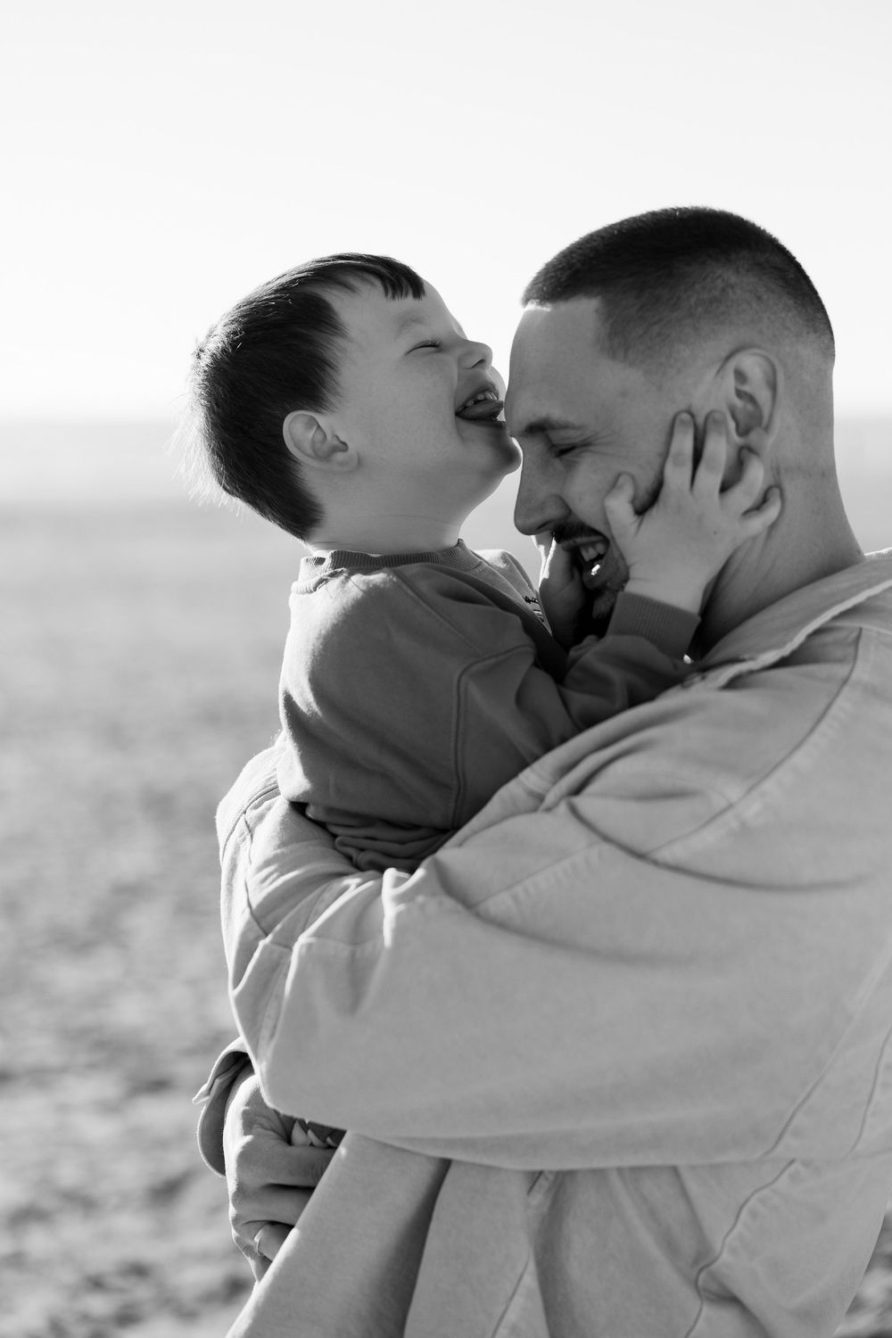 Black and white photo of a father holding his laughing young son close, sharing a tender moment outdoors.