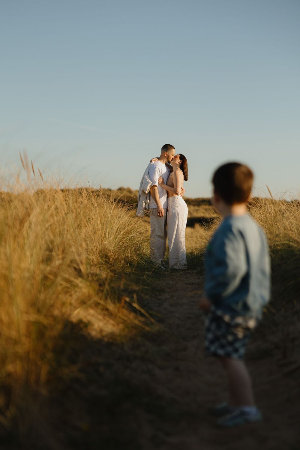 A couple shares a tender kiss on a golden field path at sunset, while a child stands in the foreground.