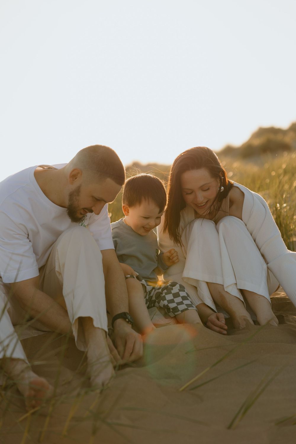 Happy family with young toddler sitting together on sunny sand dunes enjoying a golden hour outdoor moment.