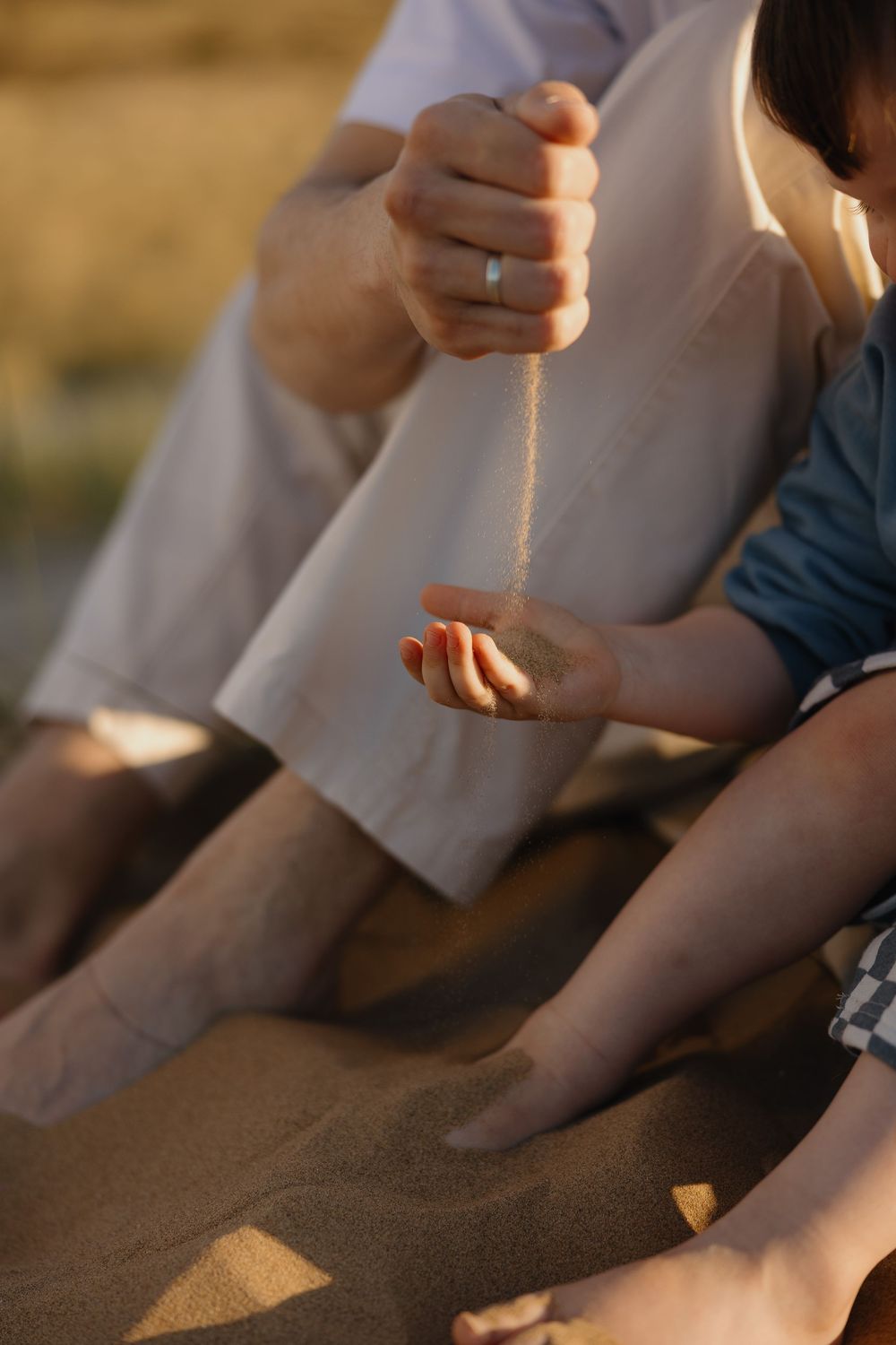 A couple sits on sandy beach, one partner pouring sand into the other's open hand in warm golden sunset light.