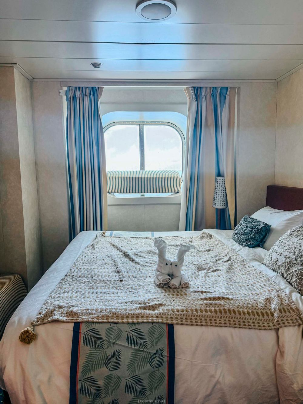Cozy cruise ship cabin with a king bed, white knit blanket, stuffed bunny, blue striped curtains, and a porthole window.