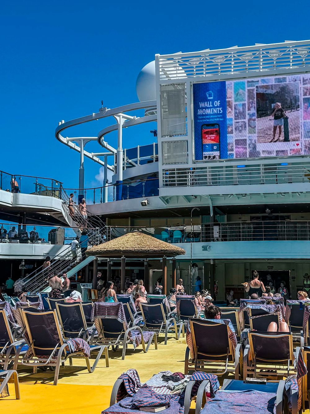 Passengers relaxing on sun deck chairs of cruise ship with water slide and large LED screen display.